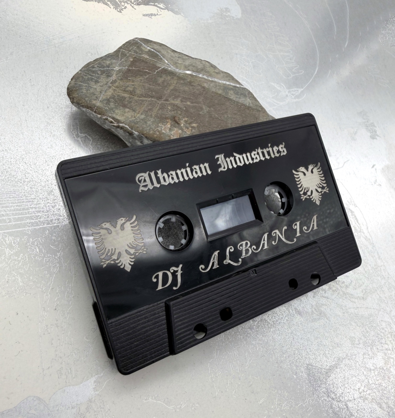 Black plastic audio cassette with pale Albanian Industries and DJ Albania lettering and two double-headed eagle motifs.